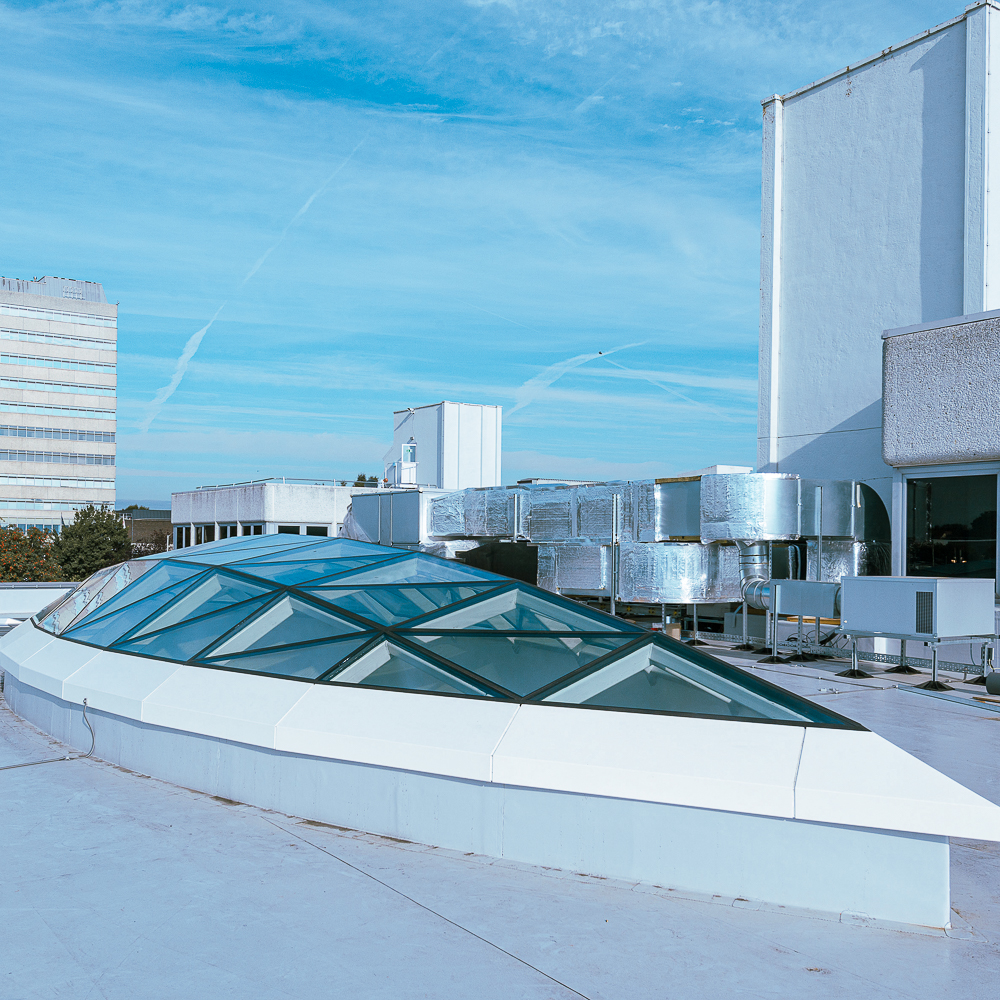 Modern rooftop with large glass panels and metallic ventilation systems against a blue sky backdrop with light clouds and contrails.