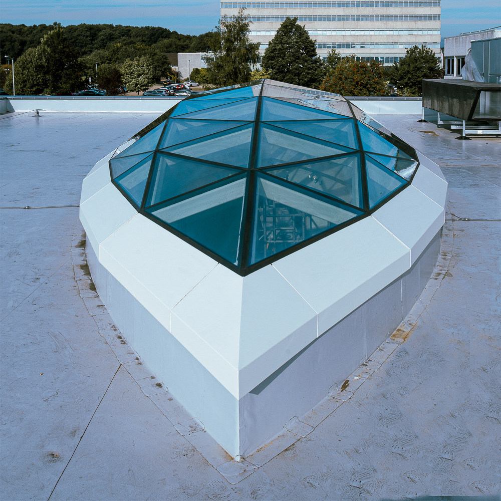 A geometric glass skylight is set in a white, angular frame on a flat rooftop. Trees and a multi-story building are visible in the background.