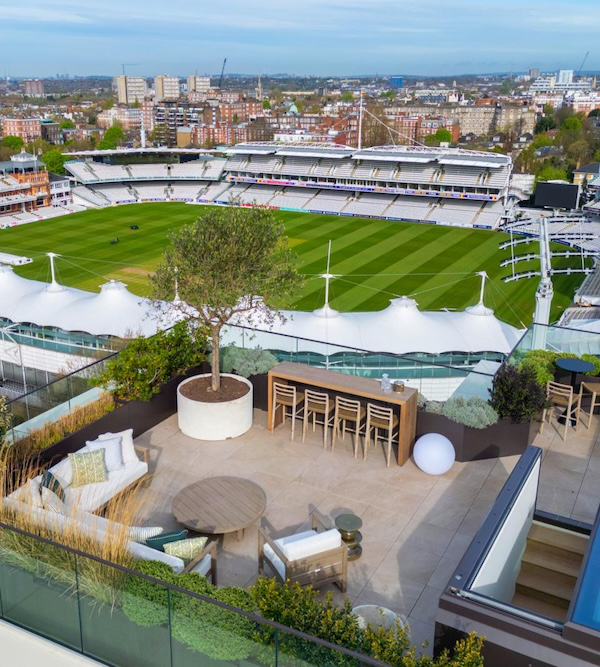 A rooftop terrace with outdoor furniture overlooks a sports stadium with a cricket field. The cityscape is visible in the background.