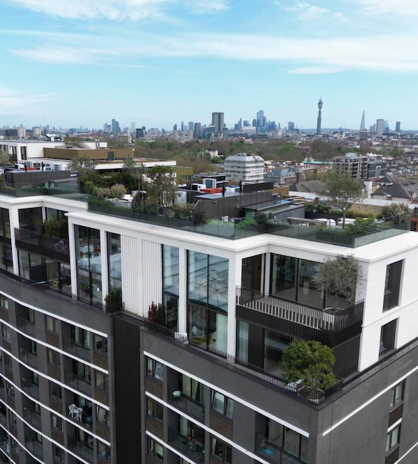 A rooftop garden and patio with trees and seating are seen atop a modern apartment building, with a city skyline featuring skyscrapers in the background under a clear blue sky.
