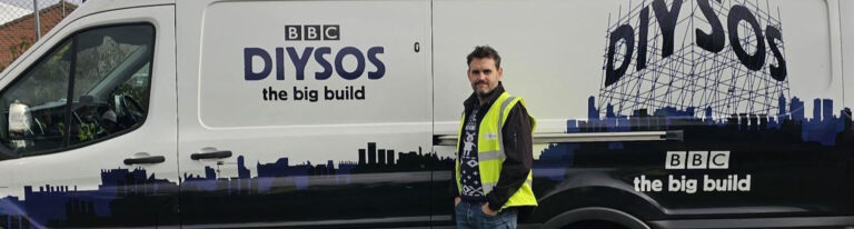 A person in a high-visibility vest stands next to a BBC DIY SOS The Big Build van with city skyline graphics.