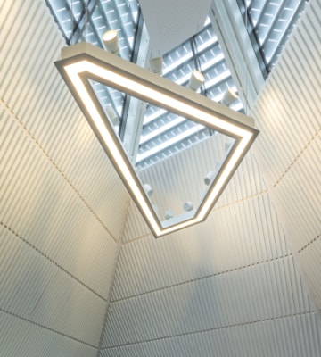 A triangular, modern light fixture hangs from a ceiling with corrugated white walls and a skylight, creating a geometric pattern.
