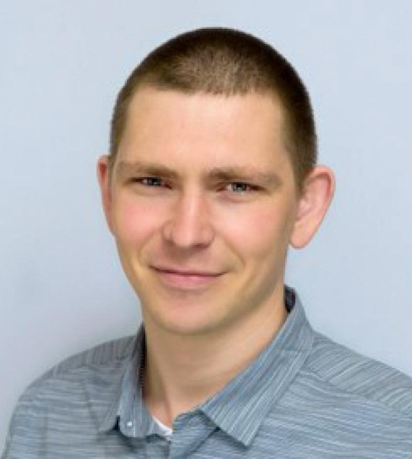 A man with short brown hair wearing a gray-striped shirt smiles in a close-up portrait against a plain background.