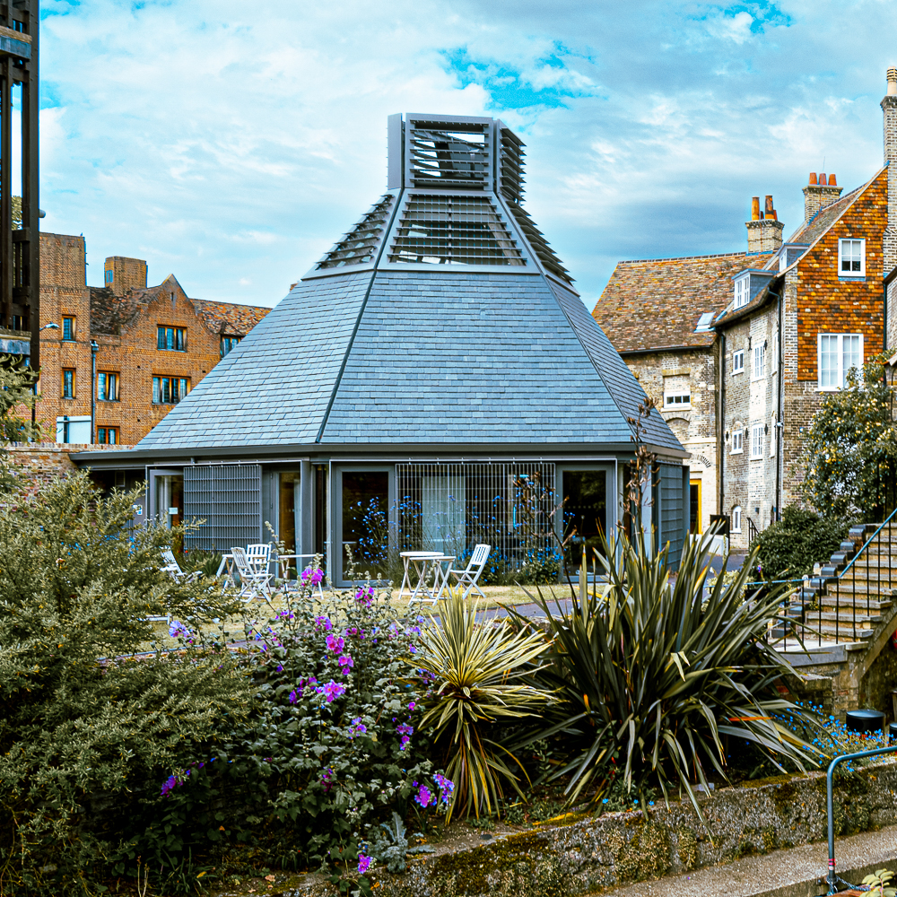 A modern, pyramid-shaped building with a slatted top, surrounded by plants and outdoor seating, stands amidst older brick buildings.