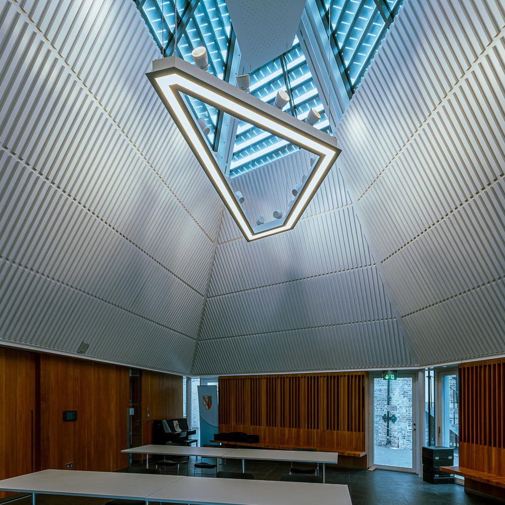 Modern triangular room with a large geometric ceiling light, wood-paneled walls, and empty tables. Skylights allow natural light in, and there's a double glass door in the background.