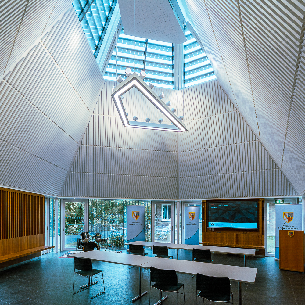 A modern conference room with a triangular ceiling, skylight, and a long table with chairs. Rectangular lights hang from the ceiling, and there are banners and a screen on the walls.