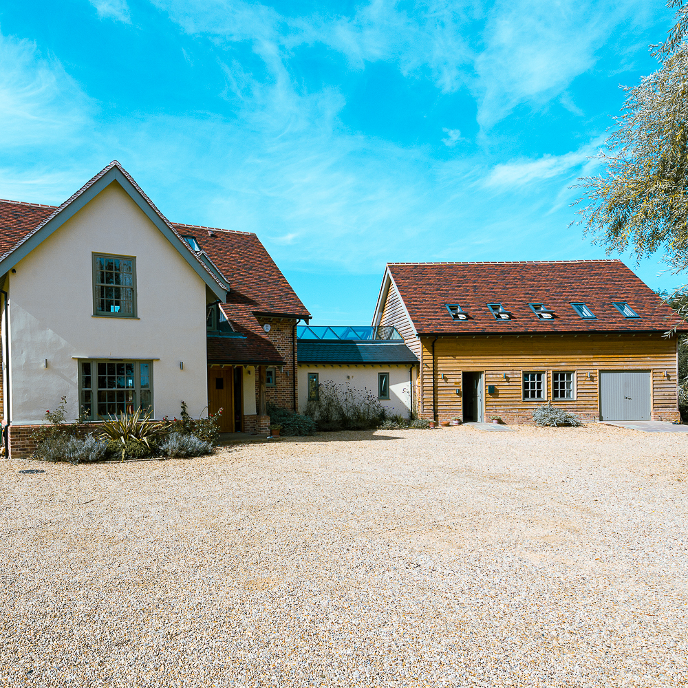 House with white walls and red roof next to a wooden structure under a clear blue sky. Gravel driveway in the foreground.