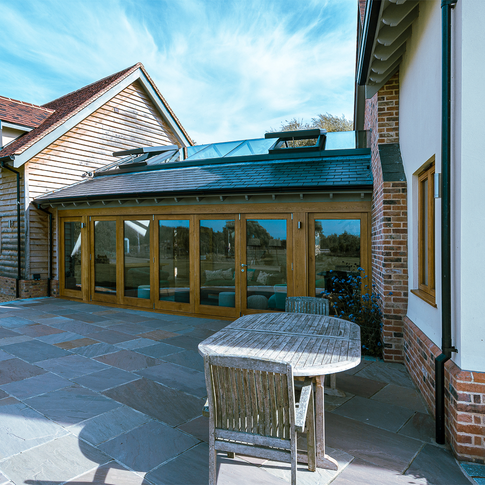 A modern house exterior with large glass doors. A wooden table with two chairs sits on a stone patio. The sky is clear and blue.