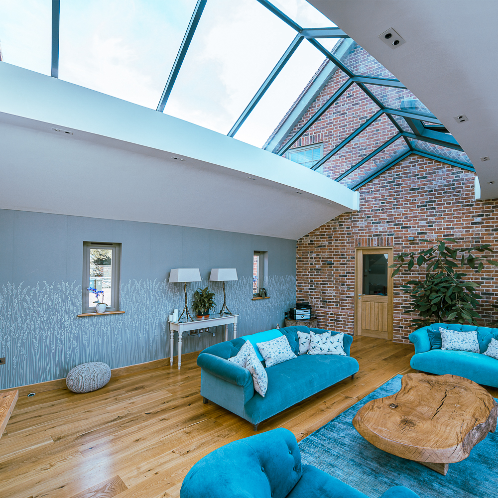 Modern living room with blue sofas and a unique wooden coffee table. Skylight ceiling and exposed brick walls add an airy feel. Decorative plants and lamps accent the space.