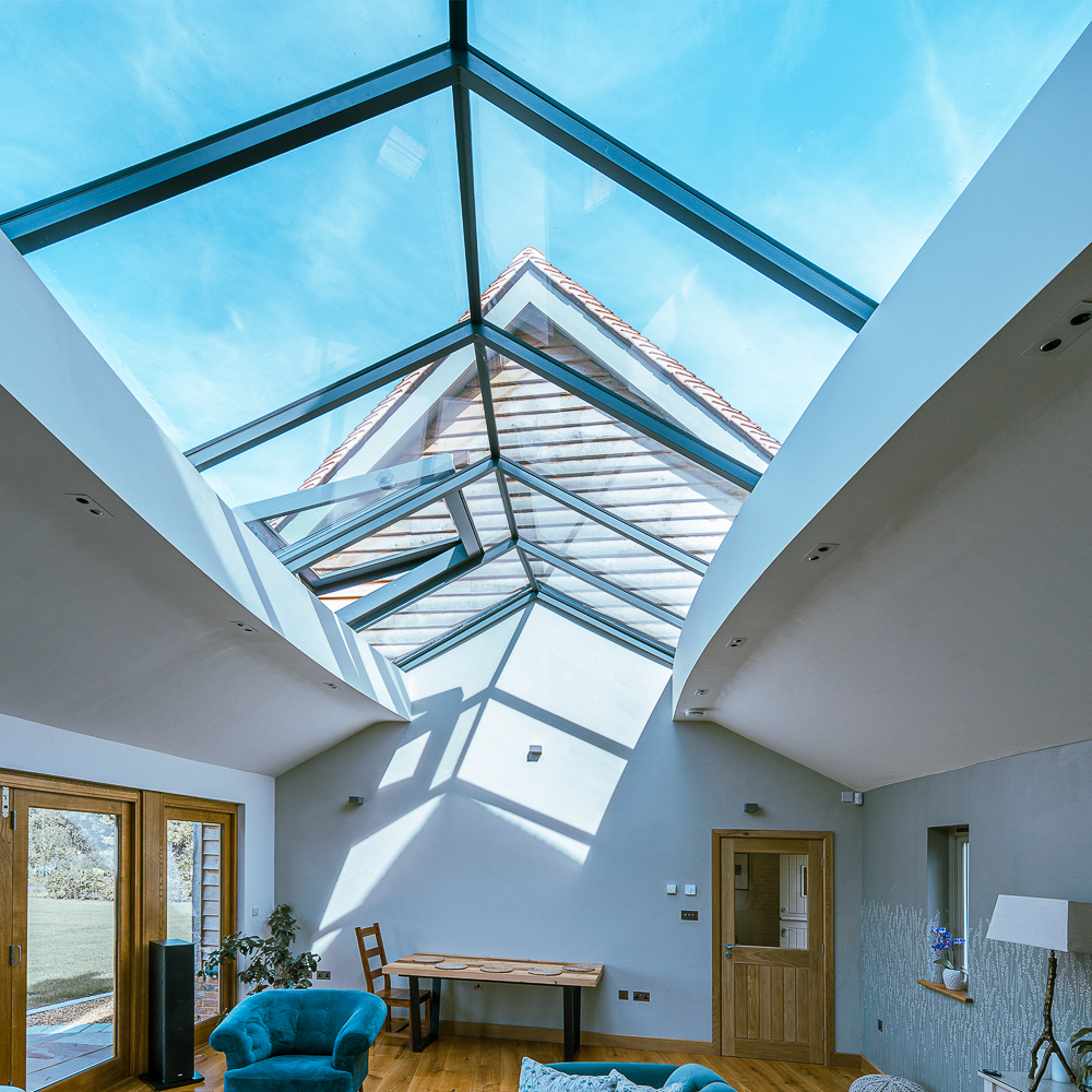 Room with a glass ceiling and wooden floor, featuring a blue armchair, a table with a chair, and wooden doors leading outside. A clear sky is visible through the glass roof.