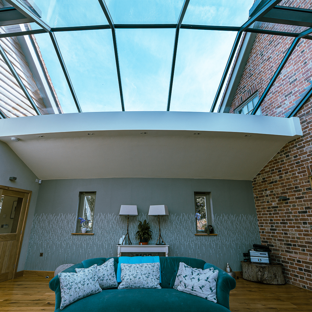 Glass-ceiling living room featuring a green sofa with patterned cushions, two lamps on a console table, and exposed brick walls.