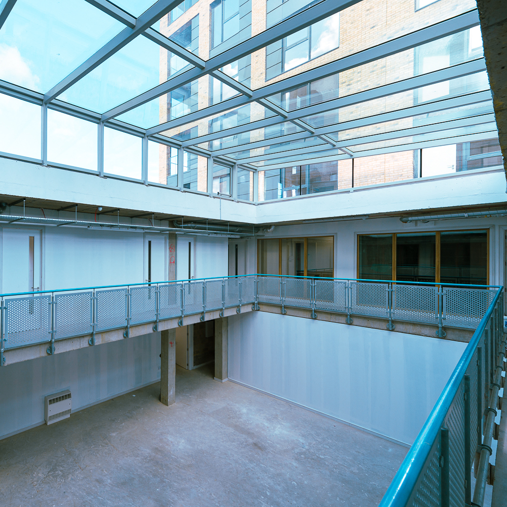 Interior view of an empty, multi-story atrium with glass ceiling and metal railings, surrounded by brick and concrete walls.