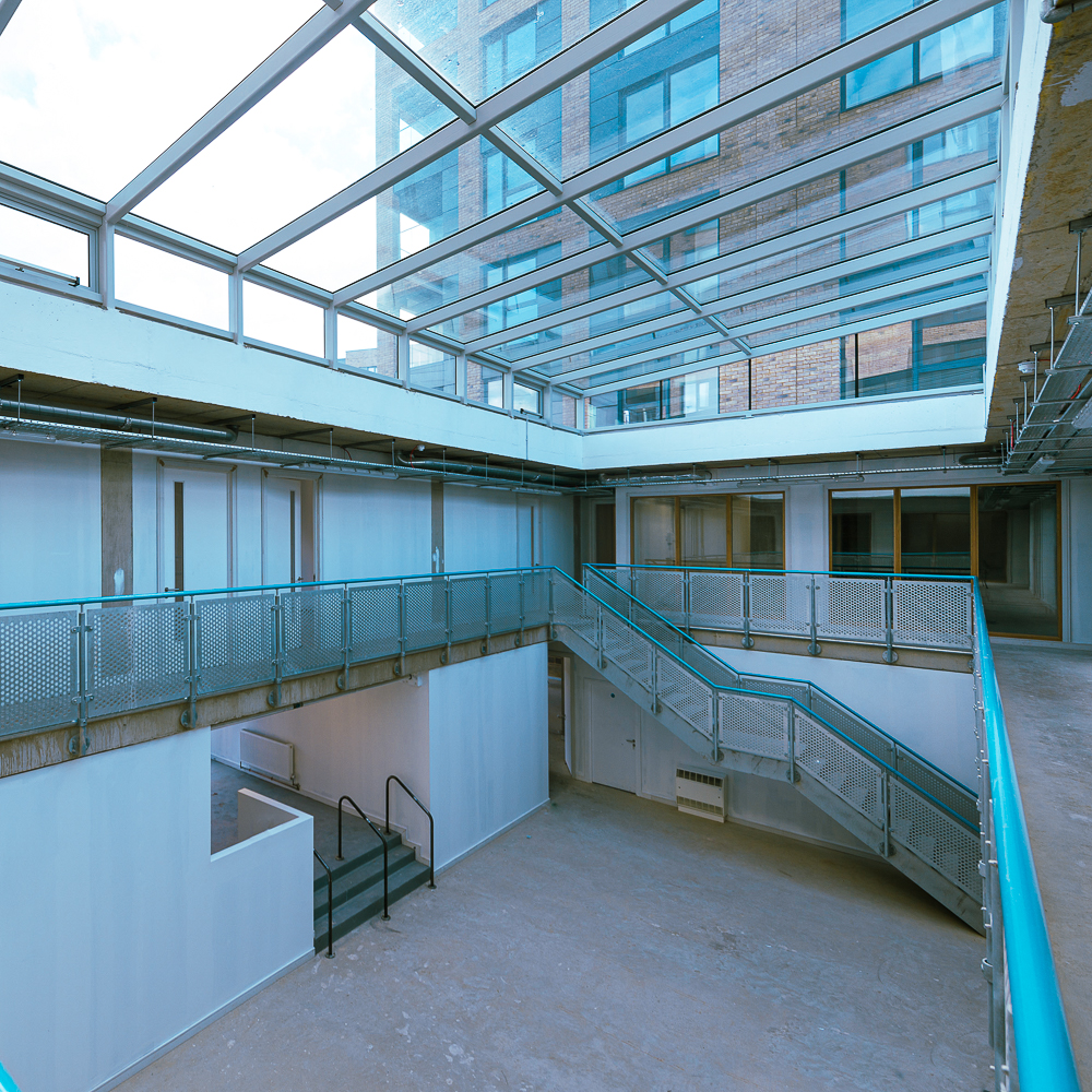 Modern indoor courtyard with a glass ceiling, featuring metal railings and a stairway, surrounded by unfinished walls and floors, in a multi-story building.