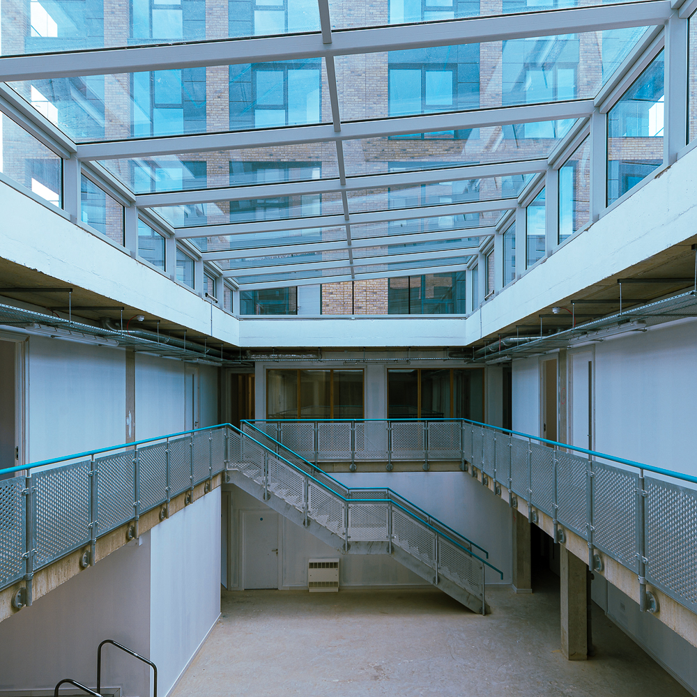 Interior of a modern building with a glass roof, featuring a metal railing and staircase, surrounded by white walls and large windows.