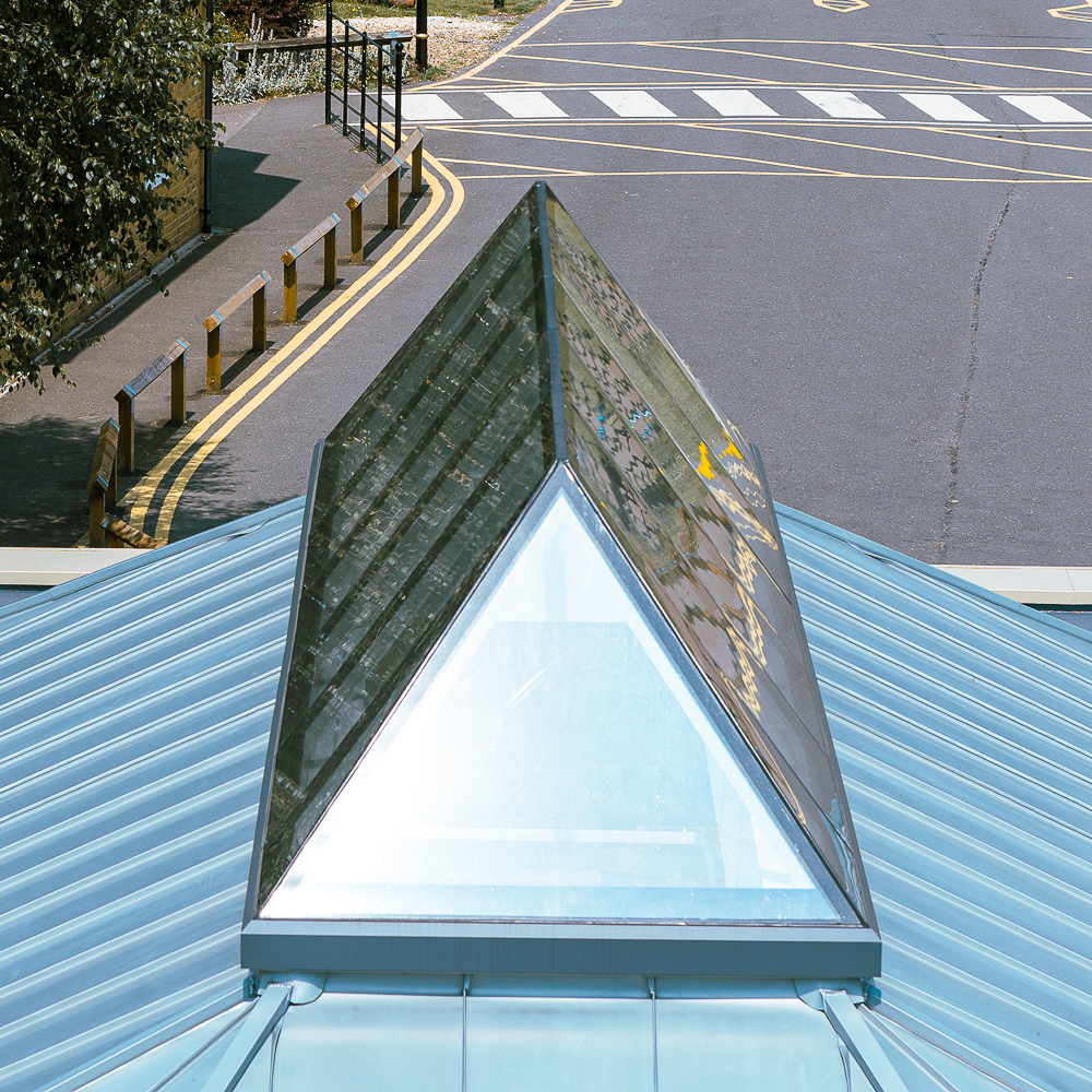 Pyramid-shaped glass roof section on a metal building, overlooking a road with painted lines and bollards.