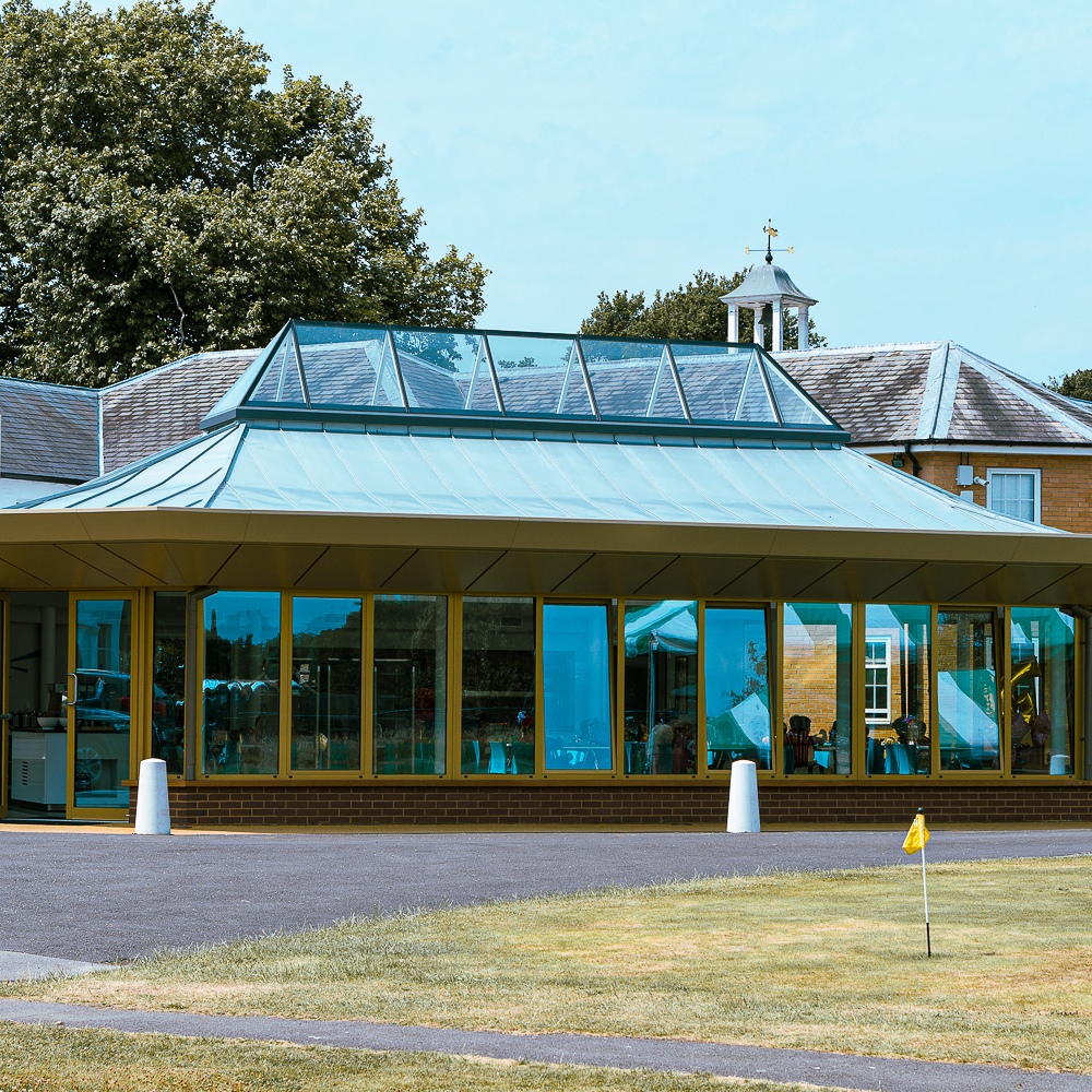 A modern glass building with a pyramid-style roof, surrounded by trees, stands near a well-maintained lawn with a small yellow flag.
