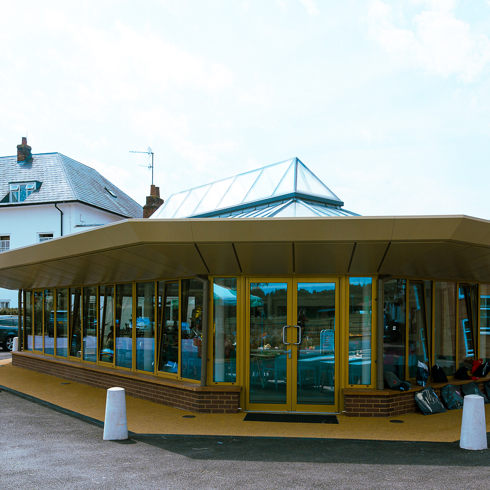 Modern building with large glass windows and a triangular glass roof. The structure features a brown and yellow exterior. Nearby, a white two-story house and vehicles are visible.