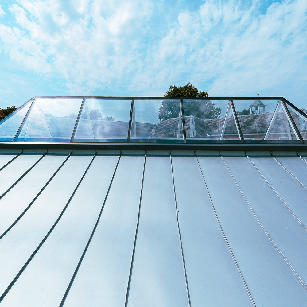 A metal roof with a row of large skylights under a blue, partly cloudy sky. Trees are visible in the background.