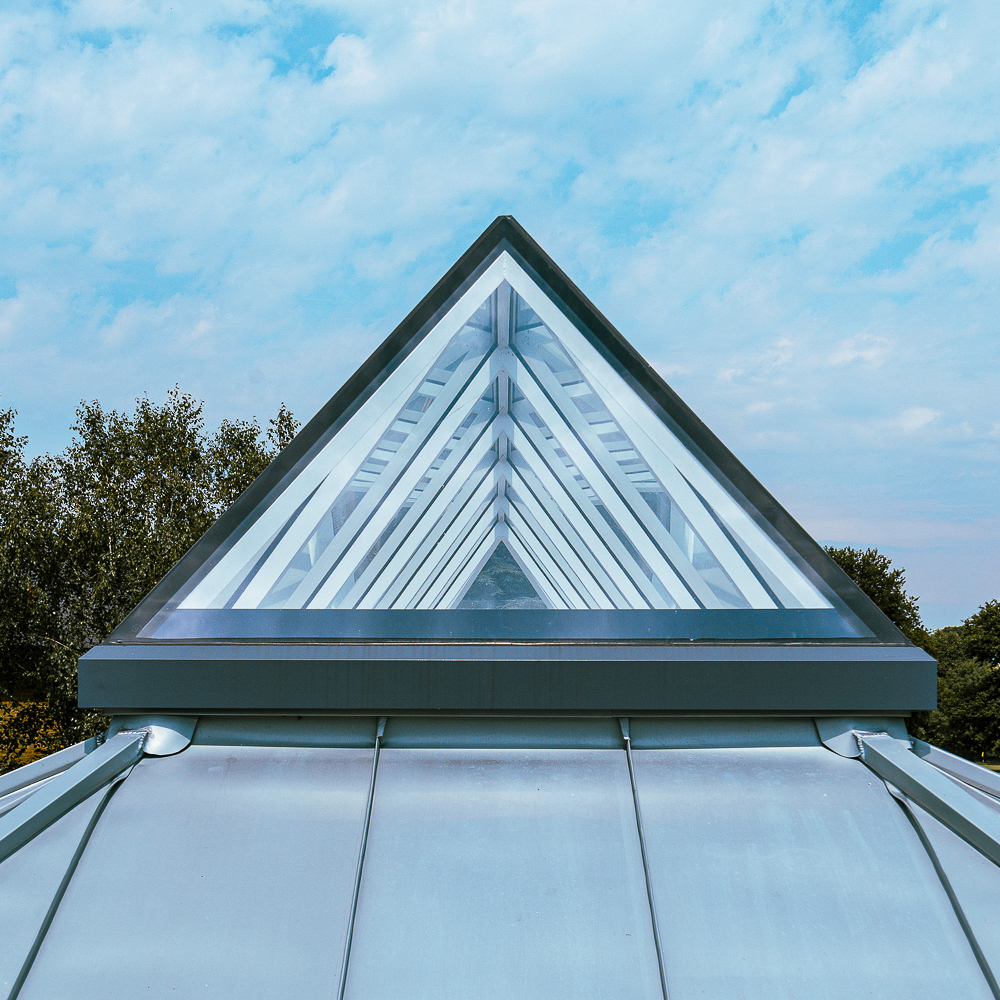 Triangular glass roof structure with metal framing, set against a blue sky and surrounded by trees.