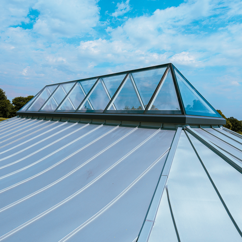 A modern building with a sloped metal roof featuring a large glass pyramid skylight, set against a blue sky with clouds.