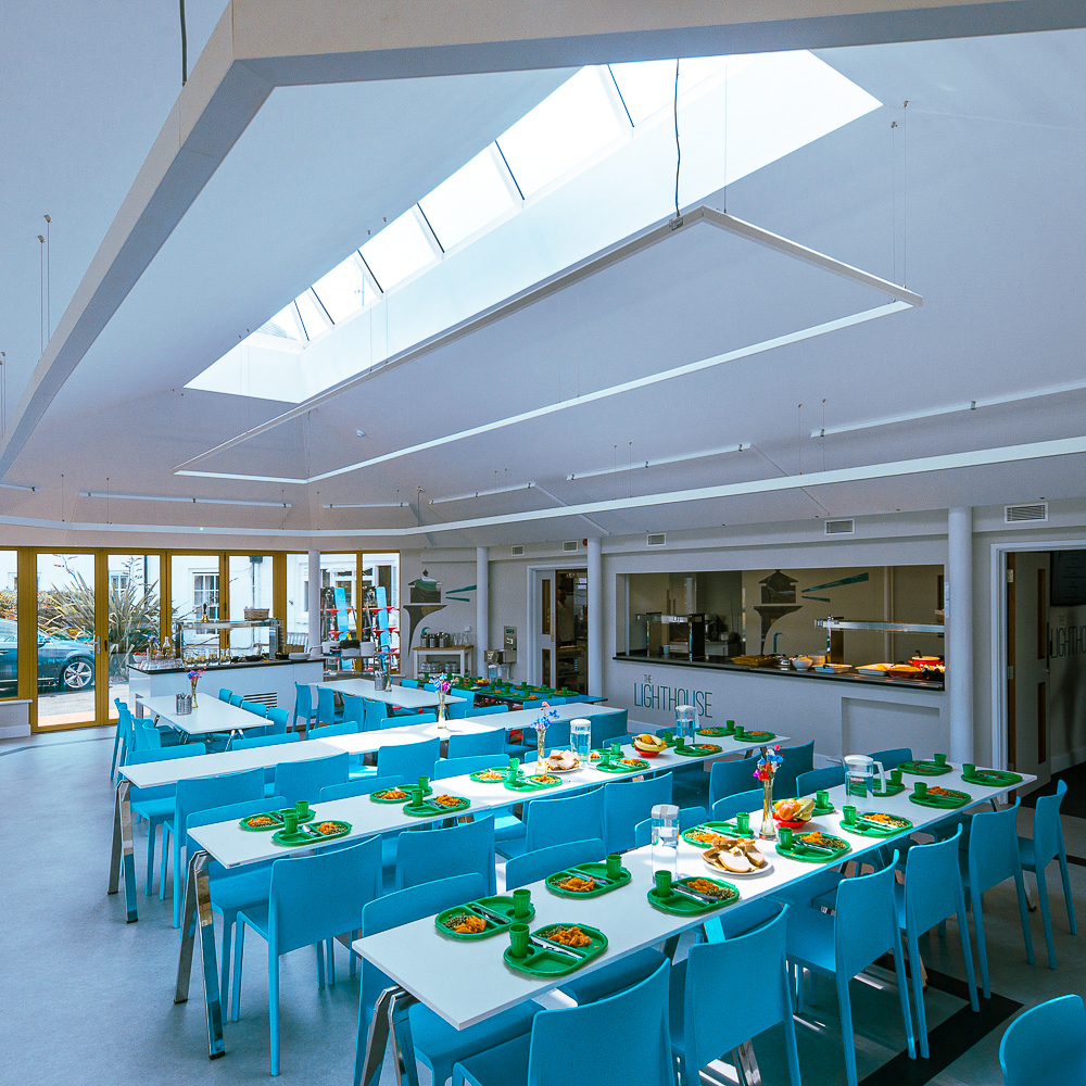 A bright cafeteria with blue tables and chairs, skylights overhead. Plates of food and trays are neatly arranged on tables.