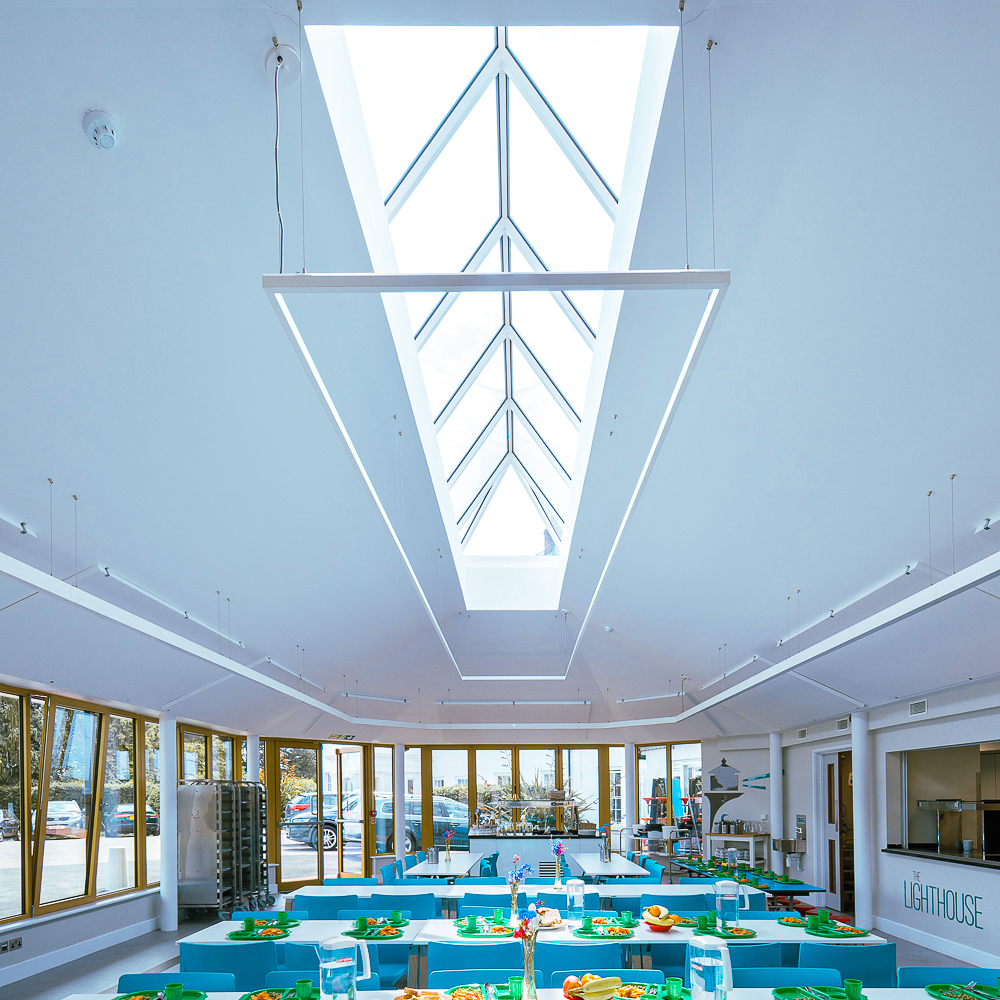 A bright, modern cafeteria with large skylight, turquoise tables, and neatly arranged food. Glass doors and windows line the walls, allowing natural light to fill the space.