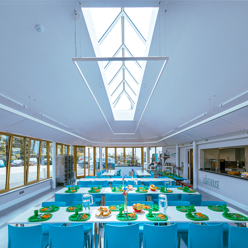 A modern cafeteria with blue tables and chairs, featuring trays of food neatly arranged. Large windows and a skylight provide ample natural light.