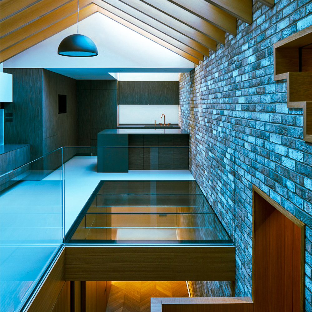 Modern interior with exposed brick wall, glass railing, and a kitchen featuring wood cabinetry. Overhead lighting illuminates the sleek design.