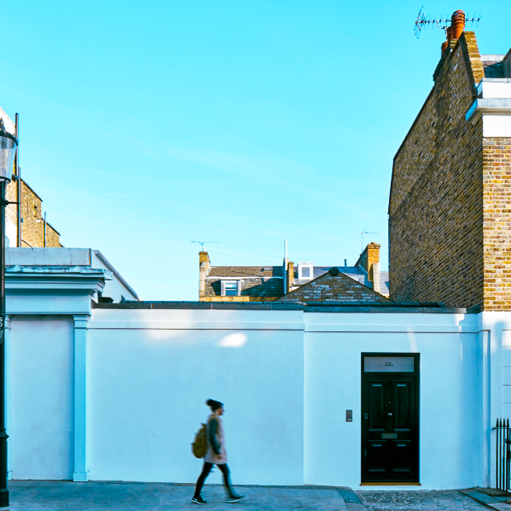 Person walking past a white wall and a brown brick building under a clear blue sky.