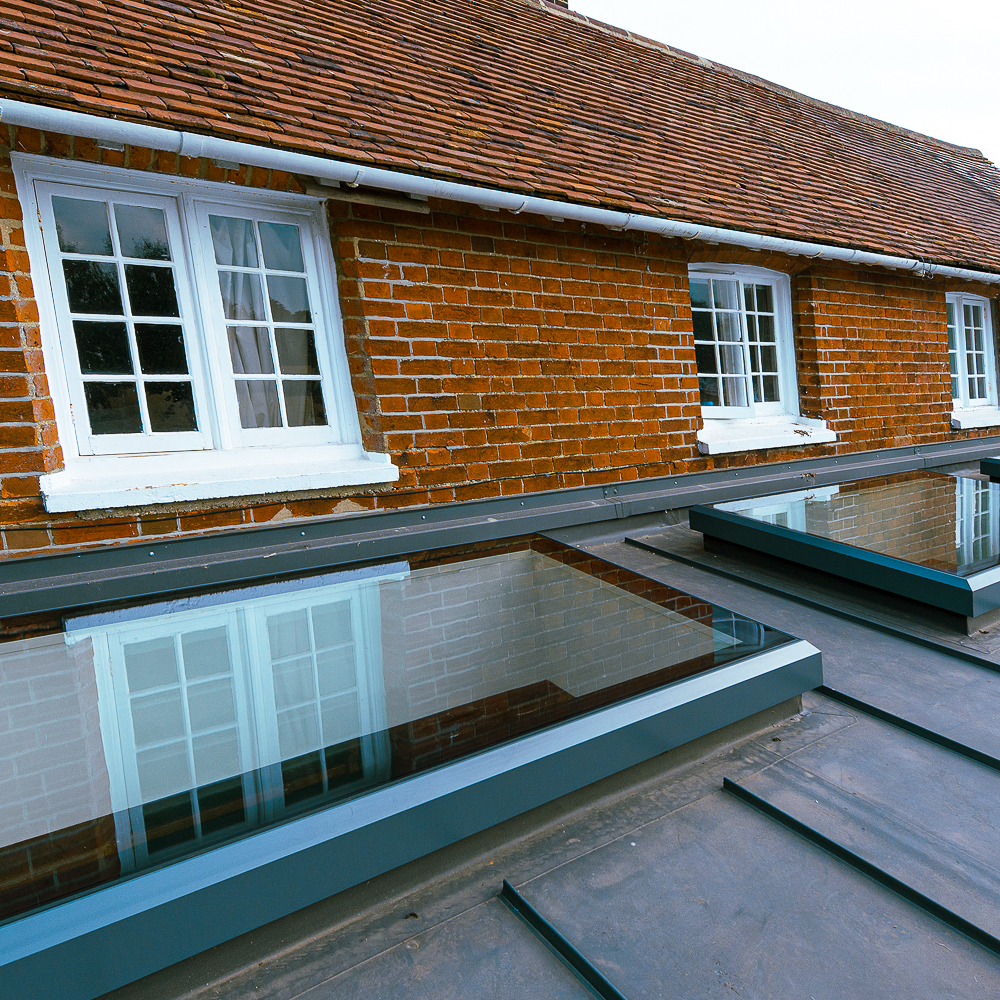 A brick house with two windows on the upper floor. Below, a flat roof features three rectangular skylights reflecting the windows.