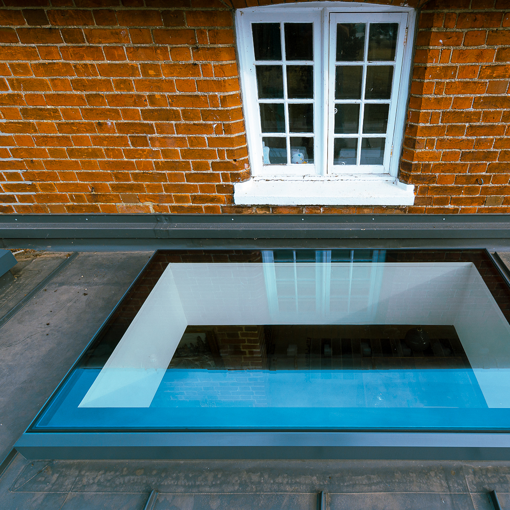 A flat skylight on a rooftop reflects a window and a section of a brick wall.