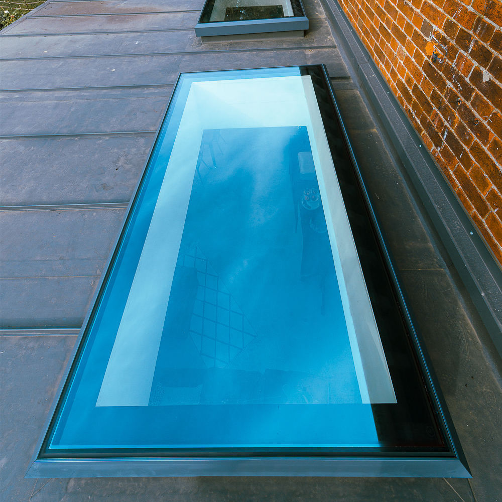 A large rectangular skylight on a flat roof reflects the sky. There is a brick wall on the right side.