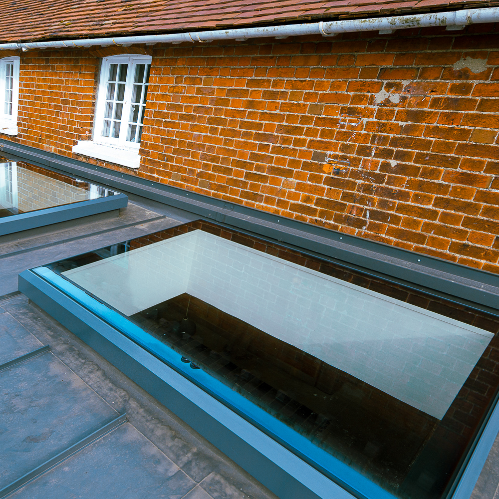 A rooftop with a rectangular glass skylight, surrounded by slate tiles, adjacent to a brick wall with two white-framed windows.