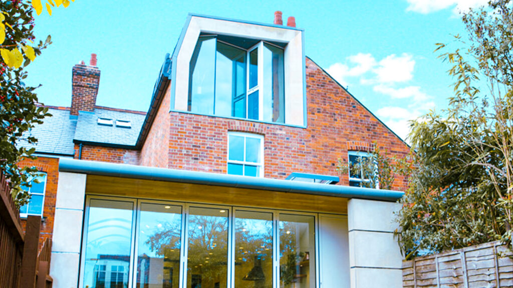 A modern house with a brick facade features large glass windows and an upper extension. Surrounding trees and a wooden fence are visible.