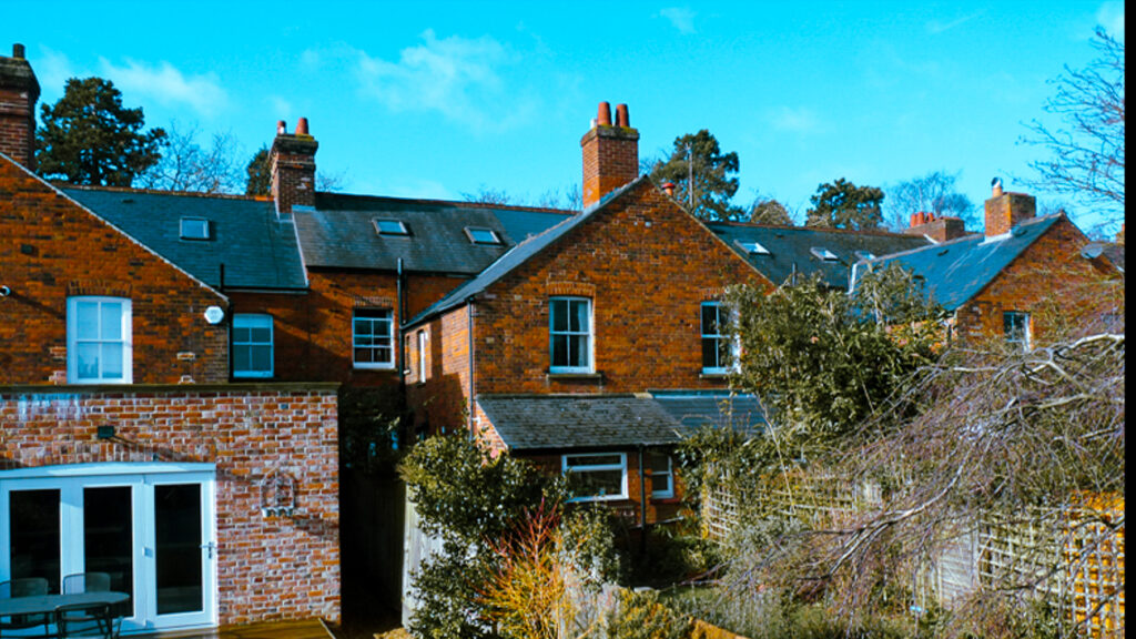 A row of red brick houses with chimneys, surrounded by trees and shrubs under a clear blue sky.