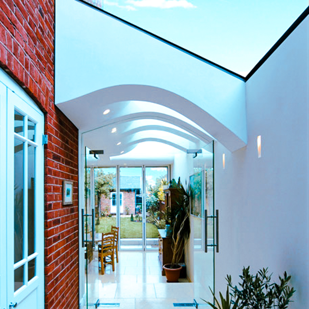 Glass doors open to a modern hallway with a curved ceiling and brick wall, leading to a bright garden space with a table and chairs.