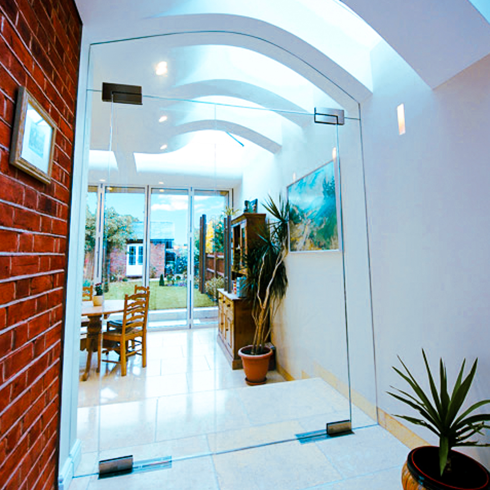 Modern hallway with glass doors, leading to a bright room with wooden furniture and plants. White walls and arched ceiling with skylights.