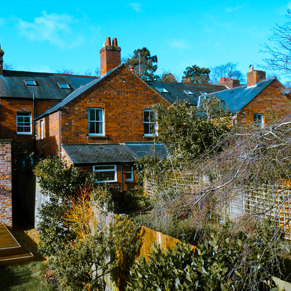 A row of brick houses with sloped roofs, surrounded by trees and shrubs under a clear blue sky.