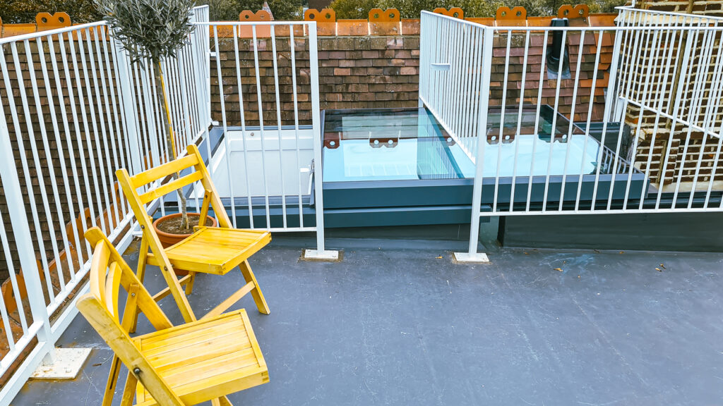 A rooftop terrace with two yellow wooden chairs and a potted plant. Glass panels are installed on the floor next to a brick wall and white metal railings.