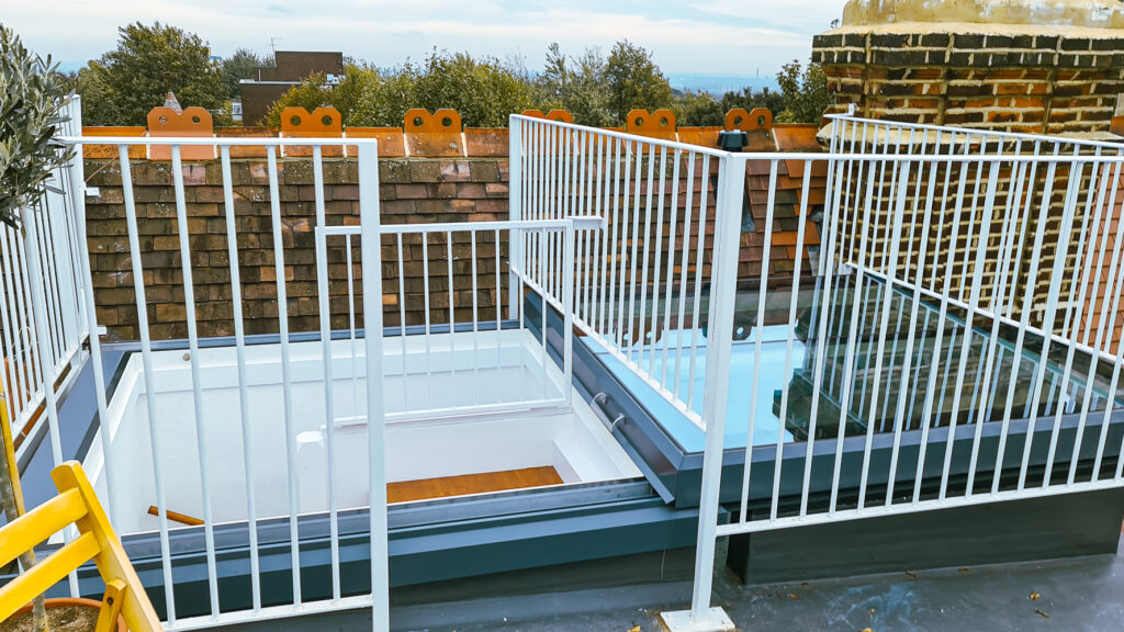 A rooftop terrace with white metal railings, a small secured gate, and skylights. Brick chimney and nearby brick wall with decorative brickwork visible in the background.