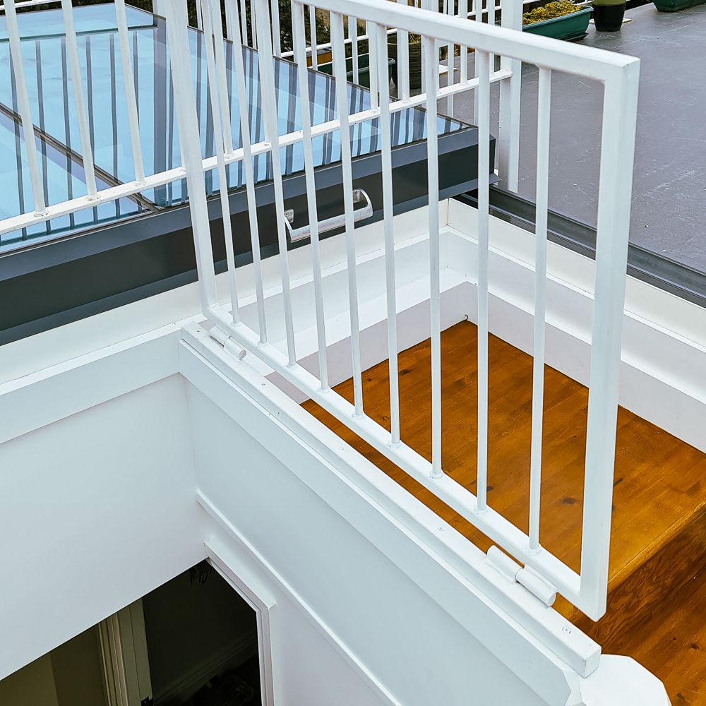 White metal railing on a staircase beside a glass roof, with a wooden floor and potted plant in the background.