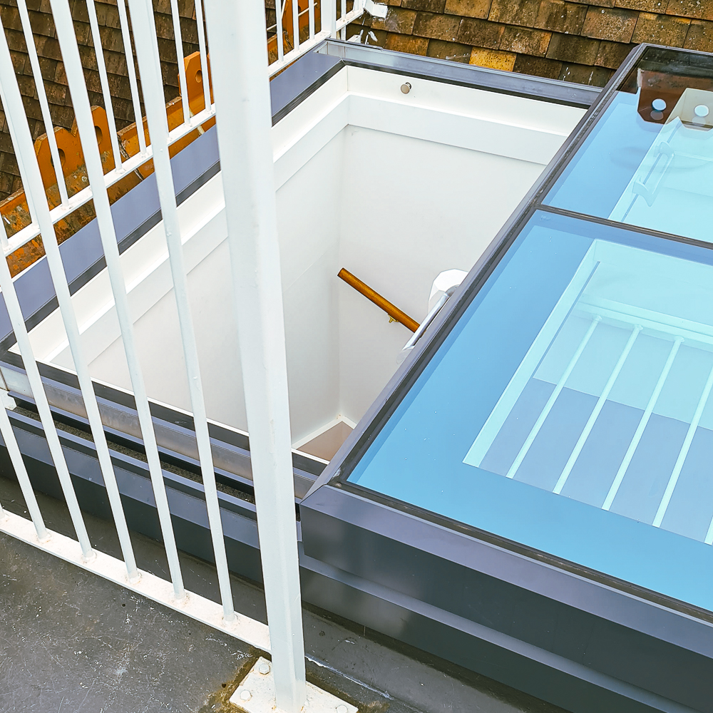A glass roof hatch with metal railing and view of a wooden handrail leading down a white staircase.