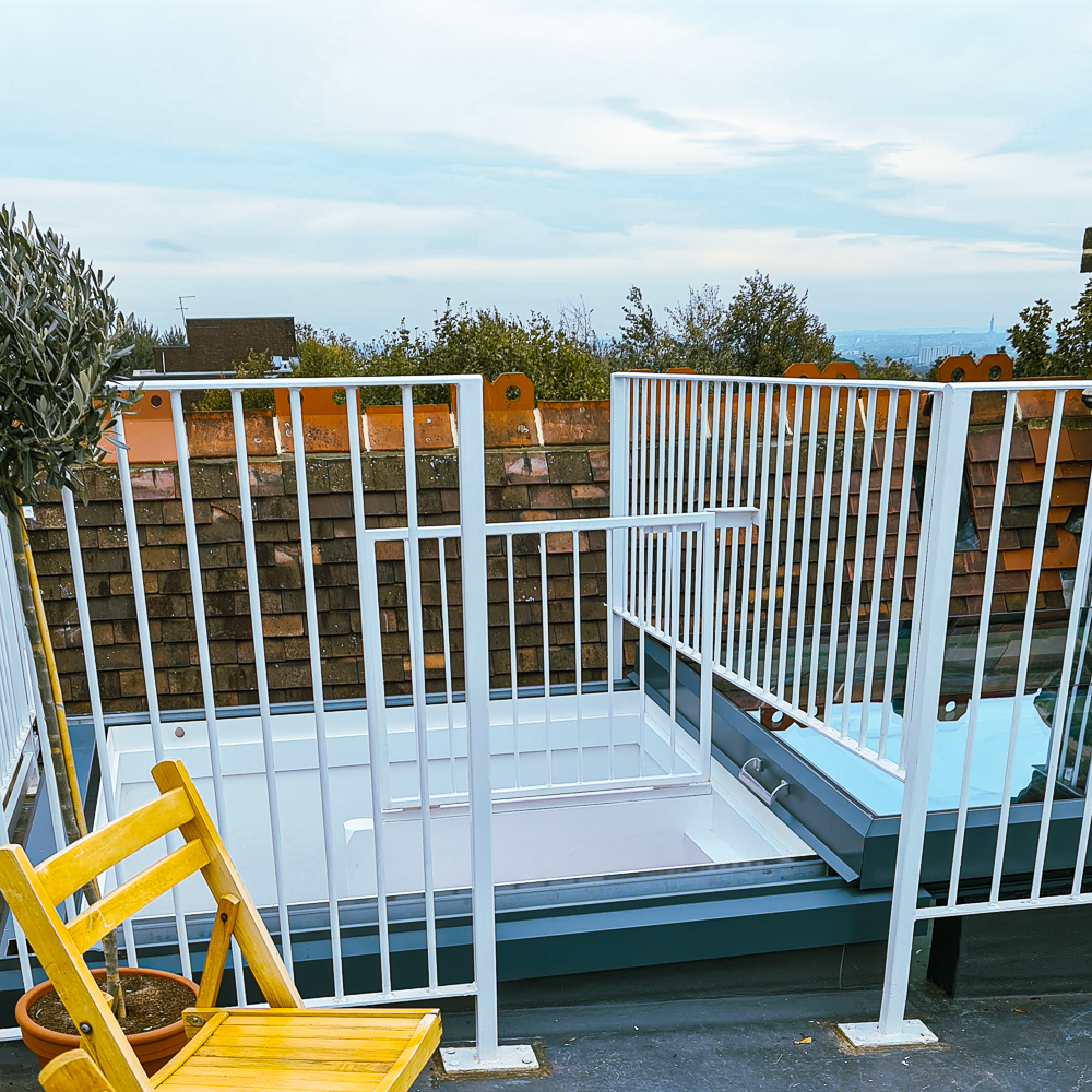 Rooftop view featuring a yellow chair, white railing, and a skylight, with a partly cloudy sky and distant landscape.