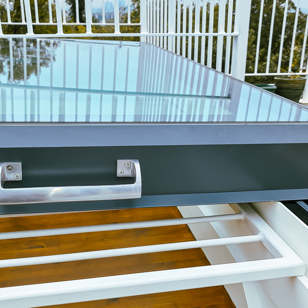A glass table with a metal handle, set on a balcony with a railing and trees in the background.
