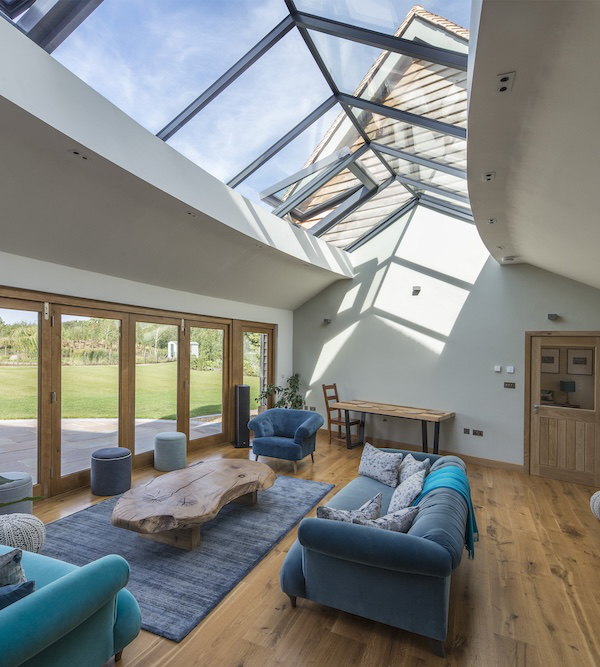 A spacious living room with large glass ceiling, wooden floor, blue sofas, a wooden coffee table, and a view of a green garden through glass doors.