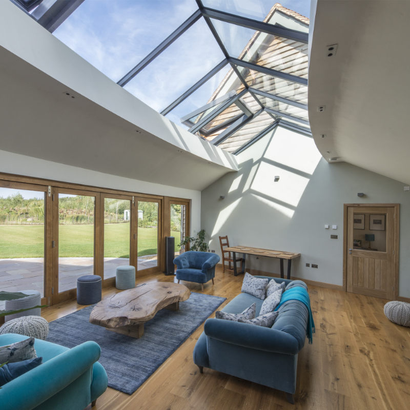 A spacious living room with a glass ceiling, wooden floors, blue and grey furniture, a wooden coffee table, and a dining table with chairs. Large windows allow natural light to illuminate the room.