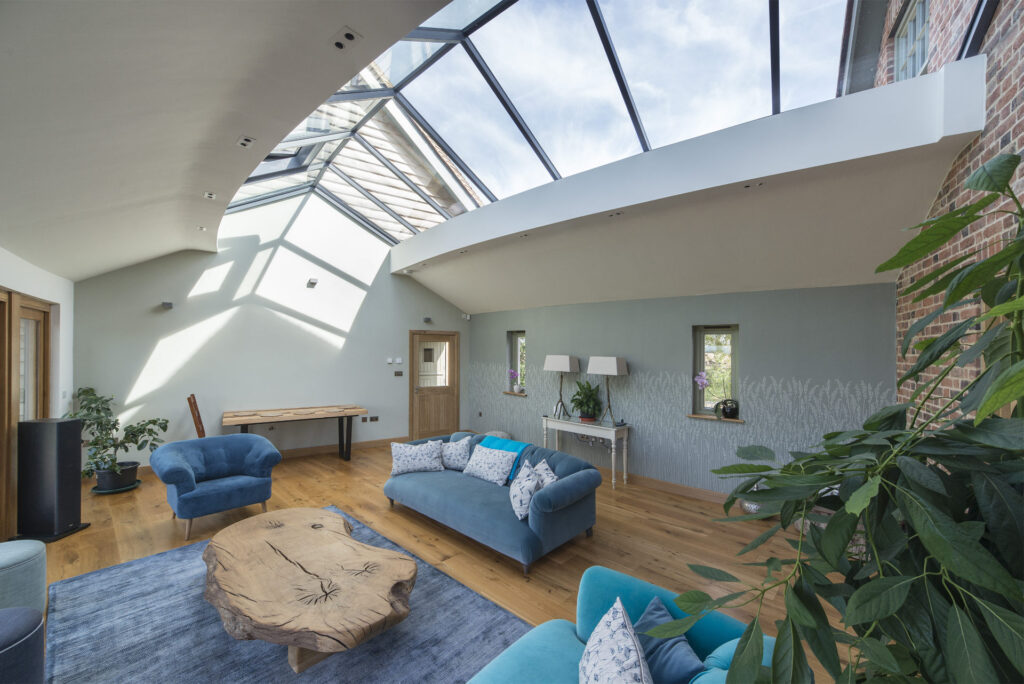 A modern living room with a glass ceiling, light blue walls, wooden floors, blue seating, a wooden coffee table, and various plants. The room is well-lit by natural light.