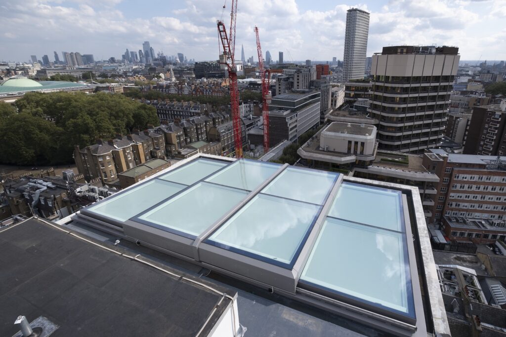 A rooftop pool with glass panels and a sliding rooflight sits atop a building, surrounded by city buildings, trees, and construction cranes under a partly cloudy sky.