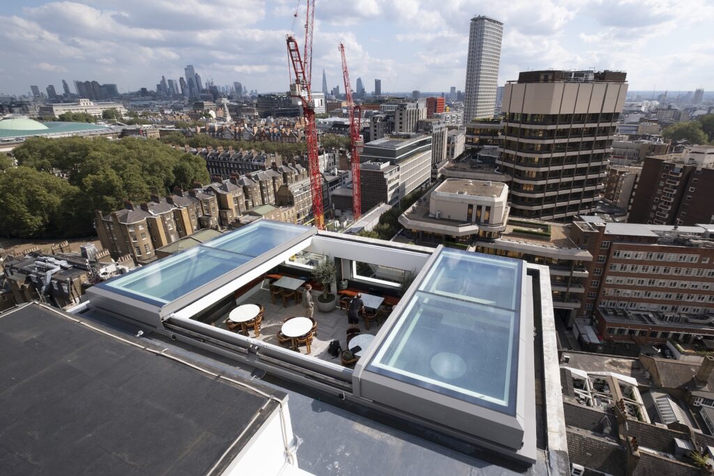A rooftop features two glass-bottomed swimming pools with seating areas between them, a sliding rooflight overhead, and is surrounded by city buildings and construction cranes under a partly cloudy sky.