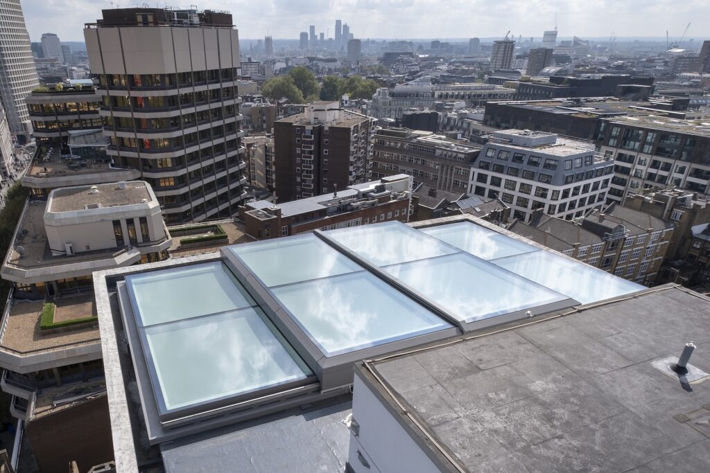 Rooftop view of a city with large sliding rooflights on a building in the foreground, surrounded by mid-rise and high-rise buildings under a partly cloudy sky.